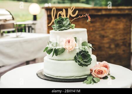 Nahaufnahme von weißen Hochzeitstorte auf dem Tisch. Love Inschrift, rosa Rose und grünen Blättern florale Dekoration. Außenterrasse. Konzept für Hochzeitstag. Stockfoto