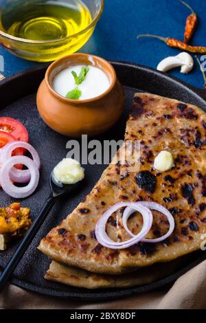 Aloo Paratha / Indische Kartoffel gefülltes Weizenbrot serviert mit Ghee (Indische Butter), Curd & Pickle, Tomate & Zwiebel. Draufsicht auf einem dunklen Moody Hintergrund. Stockfoto