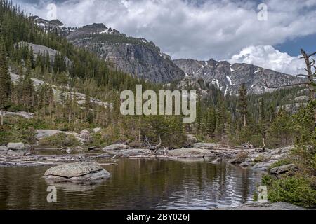 Mill's Pond Alpine Lake im Rocky Mountain National Park mit einem Felsen, Baum oder Deadfall im Vordergrund und Wolken und Bergen im Hintergrund Stockfoto