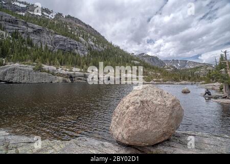 Mill's Pond Alpine Lake im Rocky Mountain National Park mit einem Felsen, Baum oder Deadfall im Vordergrund und Wolken und Bergen im Hintergrund Stockfoto