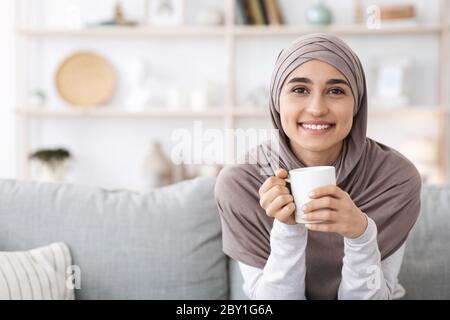 Lächelnde Arabische Frau In Hijab Auf Der Couch Mit Kaffee Entspannen, Genießen Wochenende Stockfoto