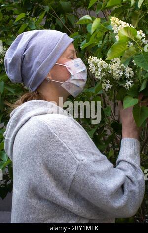 Frau in medizinischen Gesichtsmaske in grau riechenden jungen weißen lila Bund mit Knospen gekleidet Stockfoto