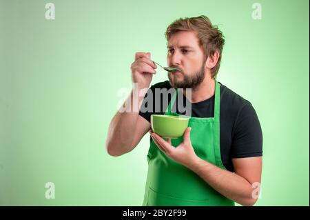 Supermarktmitarbeiter mit grüner Schürze und schwarzem T-Shirt, Suppe schmecken, Schüssel in der Hand isoliert auf grünem Hintergrund halten Stockfoto