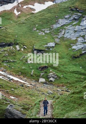 Vorderansicht des jungen Mannes mit Trekkingstöcken, der alleine in den Bergen unterwegs ist. Männlicher Wanderer zu Fuß auf dem Weg und beobachten schöne grasbewachsene Hügel. Konzept des Reisens, Wanderns und Tourismus. Stockfoto