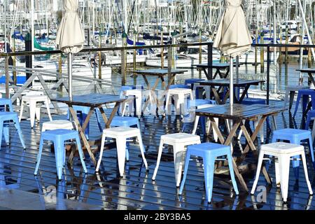 Draußen Plastikstühle und Holztische auf offener Terrasse im Yachthafen mit Liegestühlen im Hintergrund. Stockfoto