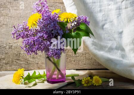 Rustikales Stillleben mit Flieder in einer Glasvase und Gänseblümchen Holzhintergrund Leinen weiß Vorhänge Nahaufnahme Stockfoto