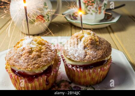 Zwei Cupcakes mit Marmelade und Sahne und einem beleuchteten Sparkler. Tasse Tee und ein Milchkännchen im Hintergrund Stockfoto