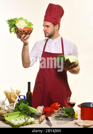 Koch steht an Tisch mit Geschirr und Zutaten. Mann mit Bart isoliert auf weißem Hintergrund. Der Küchenchef in Burgunder-Uniform hält Körbe mit Kohl und Salat. Besteck und Kochkonzept. Stockfoto