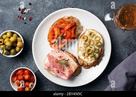 Vollkornbrot Sandwiches mit Frischkäse, Speck und Oliven Paprika in der Dose mit Tomaten auf einem Teller. Dunkler Betonhintergrund mit Kopierbereich Stockfoto