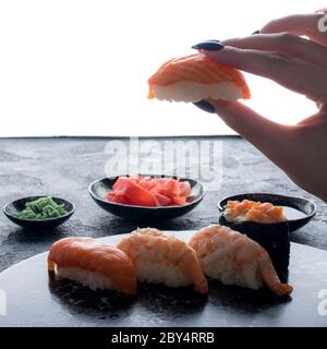 Köstliches Sushi-Set auf dunklem Steinschiefer mit Hintergrundbeleuchtung. Weibliche Hand hält leckeres Sushi Stockfoto