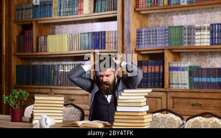 Mann in klassischem Anzug oder Professor mit gelangweilten Gesicht sitzt in der Bibliothek in der Nähe von Haufen von Büchern, Bücherregale auf Hintergrund, defokussiert. Forscher liest Buch mit gelangweilten Ausdruck. Wissenschaft und Studium Konzept Stockfoto