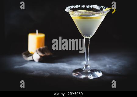 Ein Glas martini auf dunklem Hintergrund mit Zitronenschale und Salz. Duftkerze und Sticks im Hintergrund schaffen Atmosphäre. Stockfoto