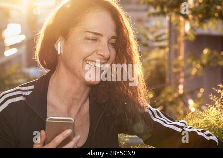 Porträt einer reifen Frau lächelnd sprechen auf einem Mobiltelefon mit drahtlosen Ohrhörern im Freien, glücklich, fit Körper Stockfoto