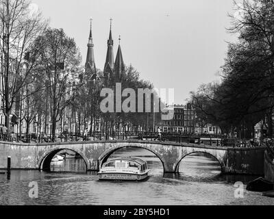 Gracht oder Canal of Amsterdam mit typischer niedriger Brücke und Sightseeing-Boot im frühen Frühling, Niederlande. Schwarzweiß-Bild. Stockfoto