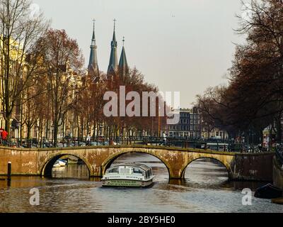 Gracht oder Canal of Amsterdam mit typischer niedriger Brücke und Sightseeing-Boot im frühen Frühling, Niederlande Stockfoto