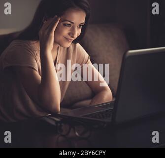 Lächelnd denkende junge schöne Frau auf Computer-Monitor und arbeiten auf dunklen Schatten zu Hause Hintergrund. Tonierter Vintage-Nahaufnahme-Zug Stockfoto
