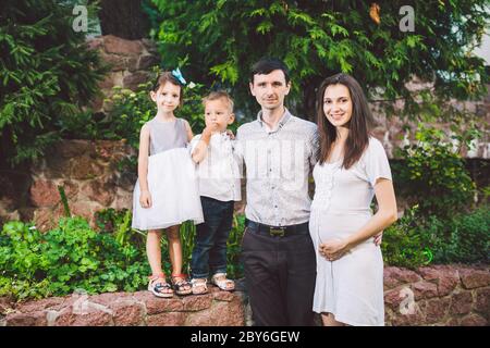 Glückliche kaukasische große Familie posiert im Hof eines Hauses im Sommer. Schwangere Frau mit zwei heterosexuellen Kindern und Familienvater stehen in der Hand Stockfoto