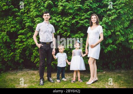 Glückliche kaukasische große Familie posiert im Hof eines Hauses im Sommer. Schwangere Frau mit zwei heterosexuellen Kindern und Familienvater stehen in der Hand Stockfoto