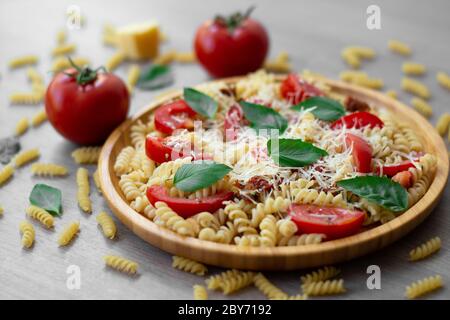 Sommerlicher kalter Nudelsalat mit Fusilli-Nudeln, frischen Tomaten, getrockneten Tomaten, geriebenem Parmesan-Käse und frischen grünen Basilikumblättern auf einem Holzteller Stockfoto