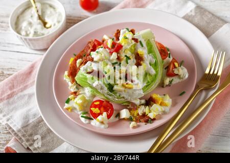 Eisberg-Keil-Salat mit Blauschimmelkäse-Dressing, knusprig gebratenem Speck, Kirschtomaten, zerbröckeltem hartgekochten Eiern, Schnittlauch auf rosa Tellern mit goldenem Schnitzel Stockfoto