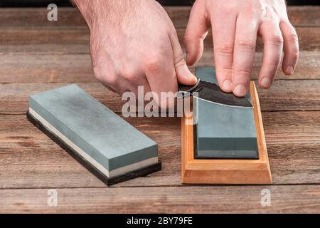 Hände des Mannes, die ein Taschenmesser mit einem Wetzstein auf einem Holztisch schärfen Stockfoto