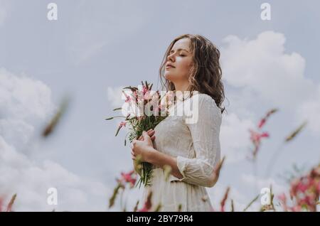 Mittelkörper Porträt einer jungen Frau in weißem Kleid mit geschlossenen Augen, hält einen Blumenstrauß, gegen blauen hellen Himmel geschossen Stockfoto
