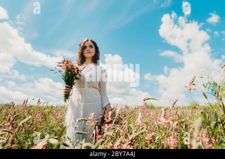 Vorderansicht Porträt einer jungen Frau in einem langen weißen Kleid stehen mit einem wilden Blumenstrauß in einem blühenden Blumenfeld gegen schönen hellen Himmel Stockfoto