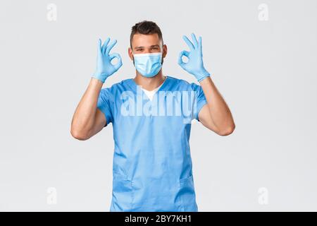 Gesundheitsarbeiter, Covid-19, Coronavirus und Prävention von Virus Konzept. Unterstützende gut aussehende männliche Krankenschwester, Arzt in Peelings zeigen okay Zeichen, Garantie Stockfoto