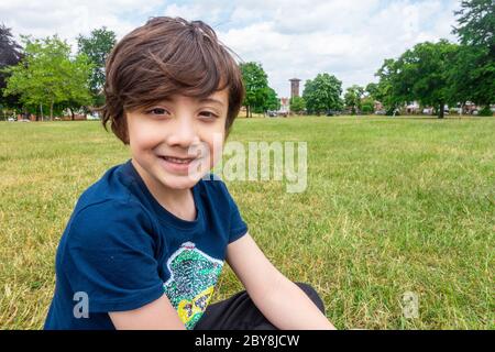 Ein kleiner Junge sitzt auf dem Boden im Park, lächelt und posiert für ein Porträt. Stockfoto