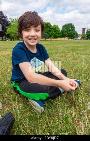 Ein kleiner Junge sitzt auf dem Boden im Park, lächelt und posiert für ein Porträt. Stockfoto