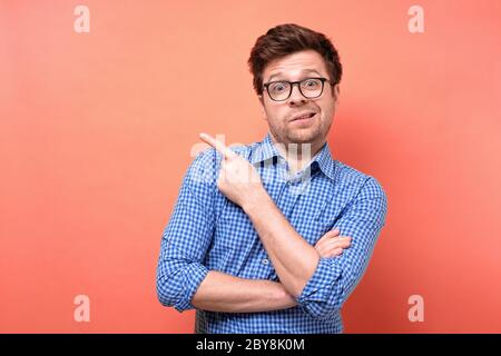 Verwirrt verwirrt verwirrt Mann in Brille und blauem Hemd Zeigen Finger beiseite isoliert über roten Hintergrund Stockfoto