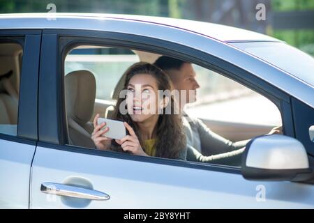 Junges Paar sitzt im Auto, Frau macht Fotos und sieht überrascht aus Stockfoto