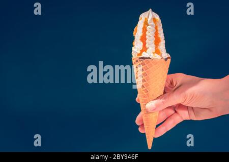 Nahaufnahme einer Hand, die einen köstlichen Vanille-Eis-Kegel mit Karamell-Belag auf blauem Hintergrund für Kopierraum hält Stockfoto