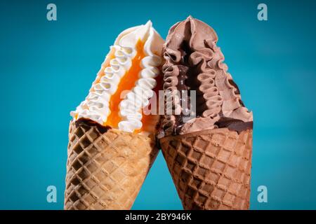 Zwei Eiscreme in Zapfen, die sich mit einem Vanillewirbel zusammenlehnen, der mit fruchtigem Sirup und Schokolade über einem blauen Hintergrund mit Vignette beträufelt ist Stockfoto