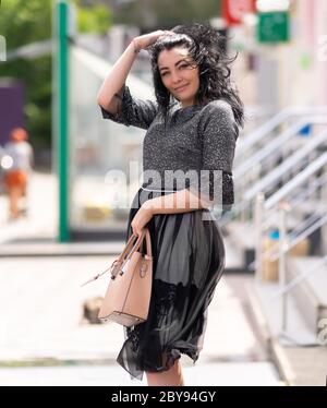 Glückliche freundliche Frau mit ihren Haaren im Wind weht ihre Hand an ihre Stirn, als sie vor städtischen Gebäuden geht mit ihrer Handtasche l Stockfoto