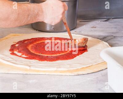 Pizza und Pizza machen Hand Putting Spreissauce auf ein Pizza aus einer Pfanne in einer kreisförmigen Bewegung Stockfoto