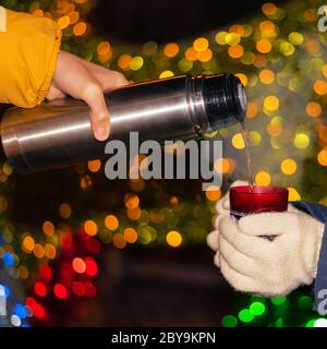 Männliche Hand Gießen heißen Kaffee oder Tee aus Thermoskannen in rote Tasse in Mädchen Hände auf dem Hintergrund der festlichen Beleuchtung. Winterurlaub Stockfoto