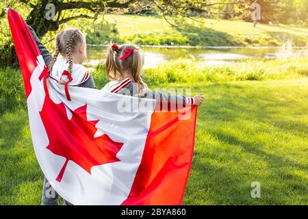 Happy Canada Day Celebration. Zwei Mädchen mit geflochtenen Haaren sind auf Naturhintergrund mit großer Kanada-Flagge in den Händen. Junge kanadische kaukasische Kinder Stockfoto