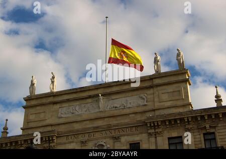 Ursprüngliches Hauptgebäude der Santander Bank (Banco de Santander) spanische Nationalflagge am Halbmast aus der Nähe von den Pereda-Gärten in Spanien Stockfoto