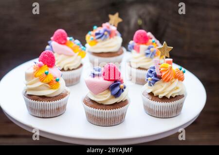 Schokoladencupcakes mit Frischkäse-Zuckerguss, pinka und Orangenkauen-Marmelade, Süßigkeiten und goldenem Topper auf dunklem Hintergrund Stockfoto