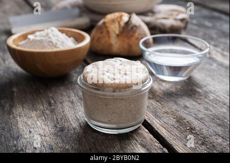 Gehäufte Dose Sauerteig Starter Hefe mit hausgemachter Brotbrötchen, Mehl und Wasser im Hintergrund. Stockfoto