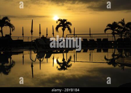 Palme silhouettiert gegen die untergehende Sonne mit Reflexion im stillen Wasser eines Schwimmbades. Keine Personen, Platz für Kopie. Stockfoto