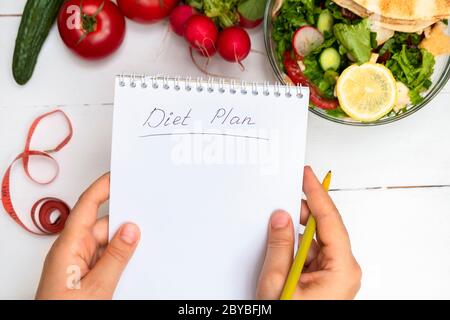 Draufsicht der Frau Hände halten Notizblock mit handgeschriebenen Worten Diät-Plan über weißem Tisch mit Gemüse und Salat Stockfoto