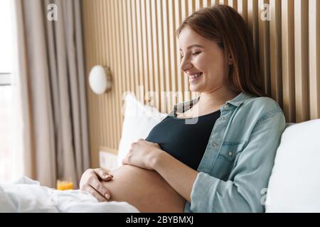 Foto von glücklicher schwangerer Frau mit roten Haaren lächelnd und streichelte ihren Bauch, während sie auf dem Bett in sonnigem Zimmer liegt Stockfoto