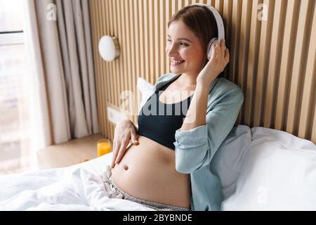 Foto von glücklicher schwangerer Frau lächelnd und mit kabellosen Kopfhörern während im Bett im sonnigen Zimmer sitzen Stockfoto