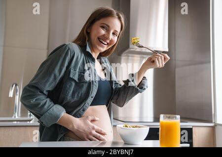 Foto von freudiger schwangerer Frau mit rotem Haar lächelnd und mit Frühstück in der modernen Küche Stockfoto