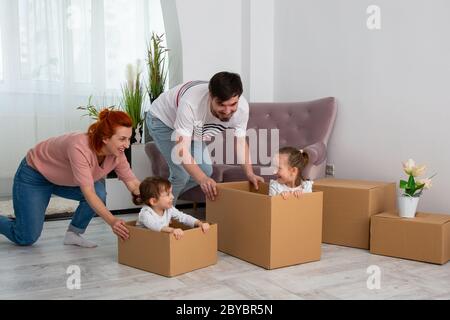 Aufgeregt glücklich junge Eltern laufen Push-Kartons mit kleinen niedlichen Kindern Töchter reiten innen, Familie Hausbesitzer spielen Spaß auf bewegen d Stockfoto