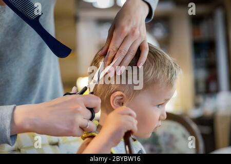 Close-up junge Erwachsene kaukasische Mutter machen Haarschnitt fot niedlich liebenswert Sohn Kleinkind Junge zu Hause durch Quarantäne und Sperrung. Mutter schneiden Haare des Kindes Stockfoto