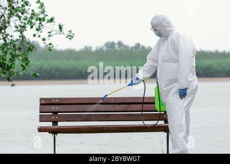 Mann trägt Schutzanzug Desinfektion der Bank mit Spray Chemikalien zur Verhinderung der Ausbreitung von Coronavirus, Pandemie in Quarantäne-Stadt. C Stockfoto