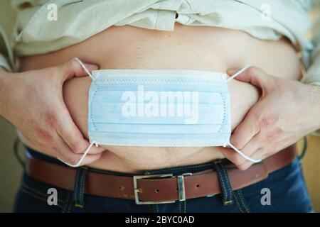 Nahaufnahme auf modernen Mann in Hemd und blaue Jeans in der modernen Haus in sonnigen Tag setzen medizinische Maske auf den Bauch. Unzufrieden mit Gewichtszunahme auf Coronavir Stockfoto